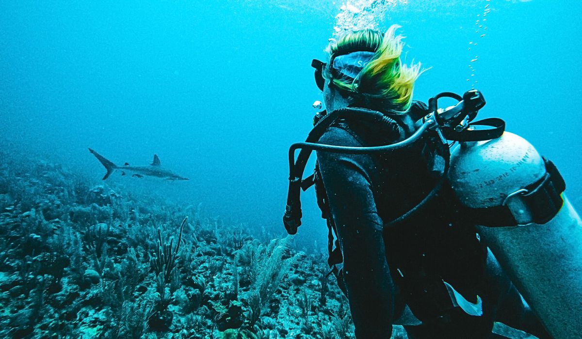 Katie scuba diving in the clear blue waters of the Great Barrier Reef near Caye Caulker, Belize, with a shark visible in the distance among the coral reef.