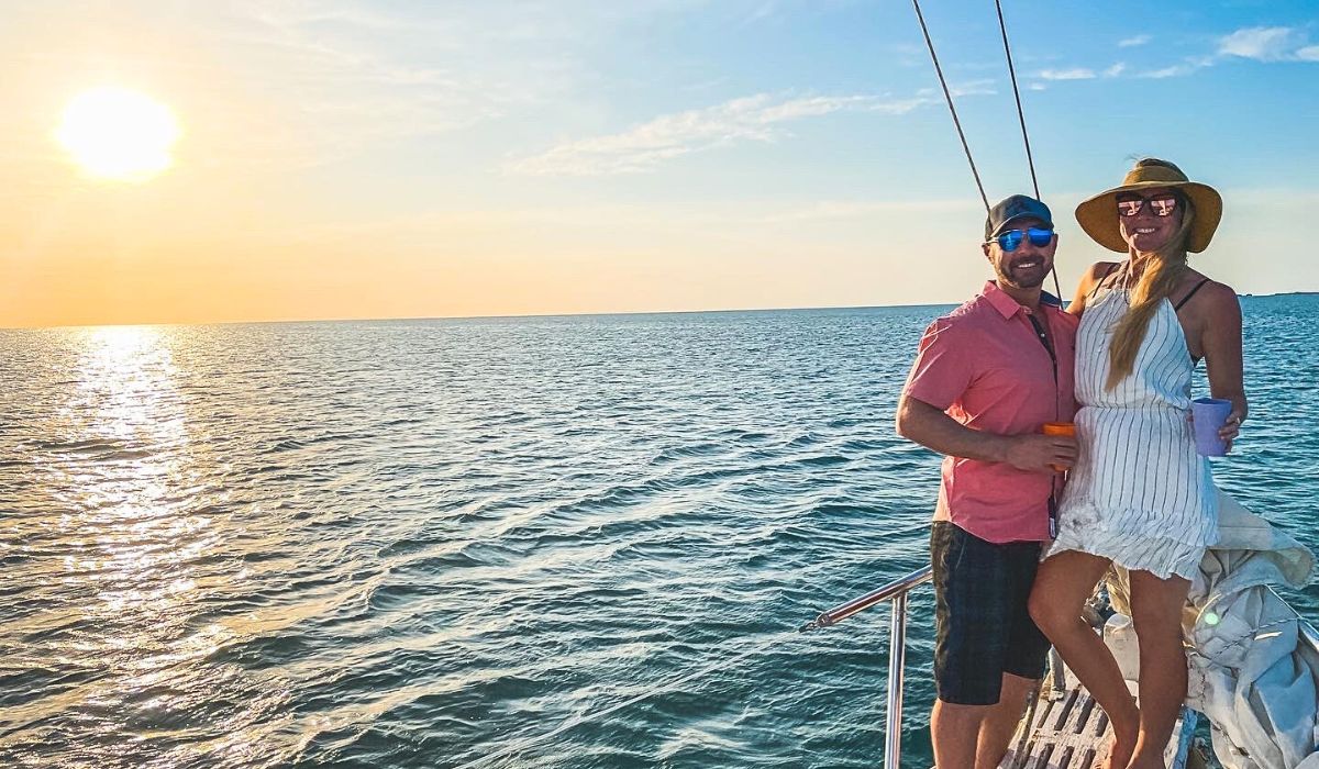 Kate and her husband enjoying a sunset cruise near Caye Caulker, Belize, standing on a sailboat deck with the sun setting over the calm ocean waters. They are smiling, holding drinks, and dressed in casual summer attire.