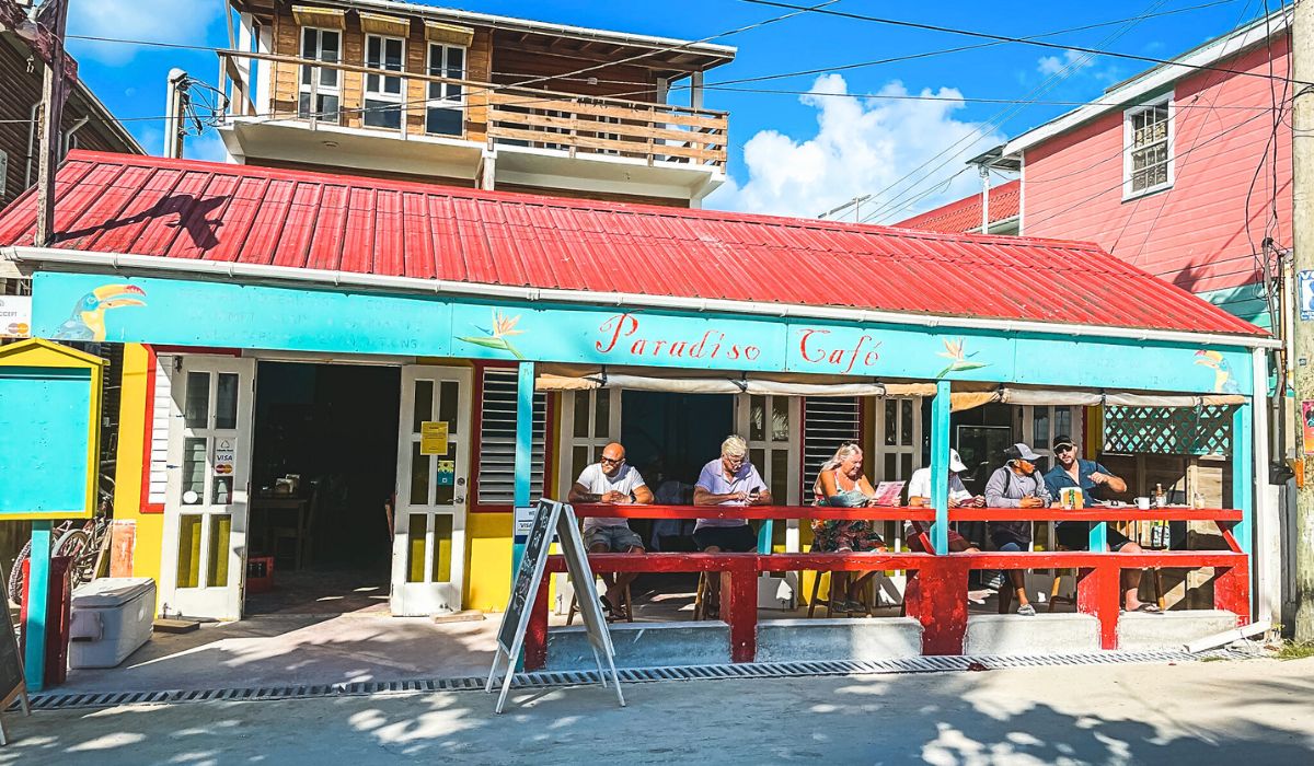 Patrons enjoying a meal at Paradiso Cafe, a vibrant restaurant with a red roof and turquoise facade on Caye Caulker, Belize. The outdoor seating area features colorful decor and a relaxed atmosphere under a sunny sky.