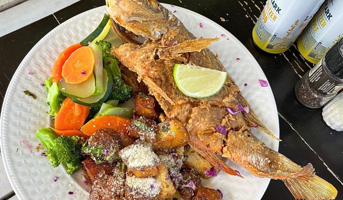 A plate of whole fried snapper served with roasted potatoes and a medley of steamed vegetables, garnished with a lime wedge at Pelican Sunset Bar in Caye Caulker, Belize. The colorful dish is complemented by cans of sparkling water and seasonings on the side.