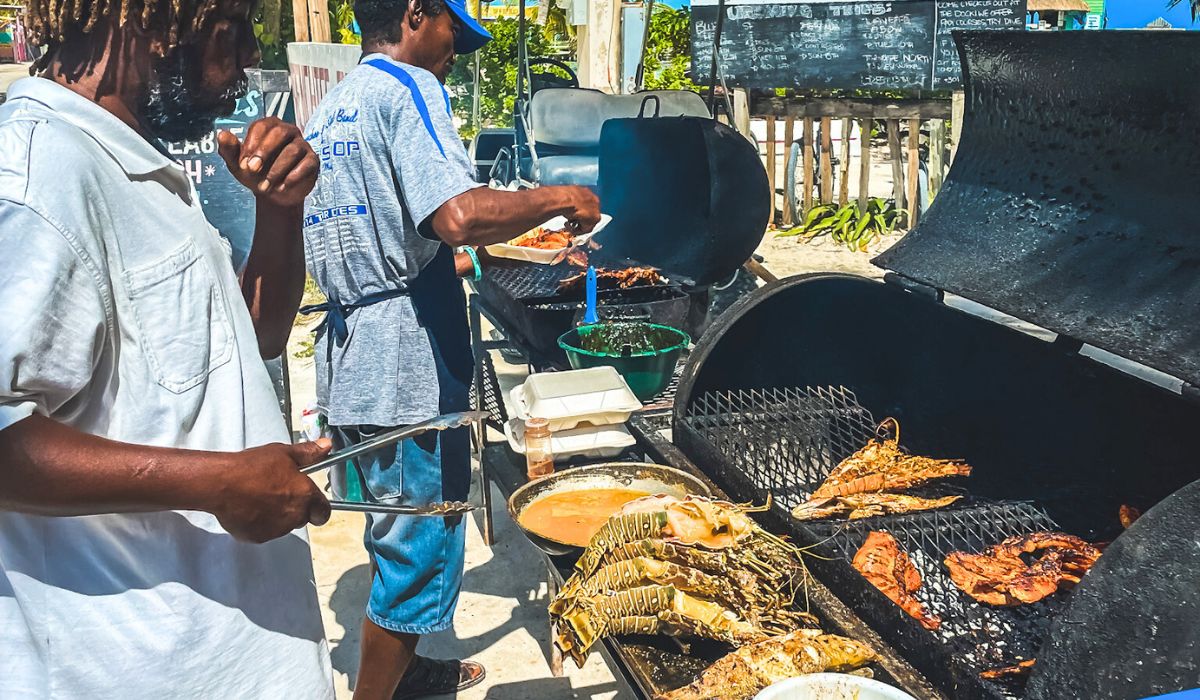 Chef Kareem grilling fresh seafood and meats on large outdoor grills for his "Unbelizeable" lunch street food in Caye Caulker, Belize. The scene captures the lively atmosphere with tantalizing food being prepared under the sunny sky.