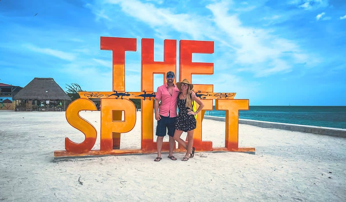 Kate from Kates Crossing Blog and her husband stand in front of a large, colorful "THE SPLIT" sign on a sandy beach in Caye Caulker, with the ocean and a blue sky in the background. This image is the cover photo for the blog post on Things to do in Caye Caulker.