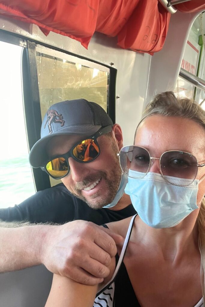 Kate and her husband wearing masks and sunglasses, sitting next to a window on the water taxi. They are smiling and enjoying the view outside while trying to stay cool inside the hot cabin.