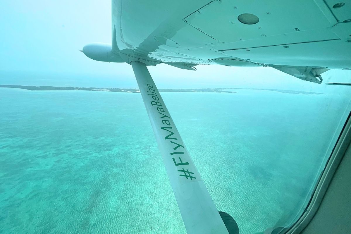 Aerial view from a small plane over turquoise waters near Belize, with the hashtag #FlyMayaBelize visible on the wing strut. The coastline is seen in the distance under a hazy sky.