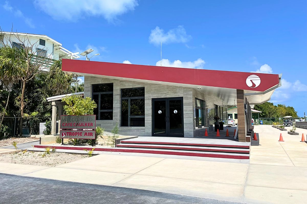 The Tropic Air terminal building in Caye Caulker, Belize, with a sign that reads "Welcome to Caye Caulker." The building has a modern design with large windows and a red roof, and the area is surrounded by tropical plants under a clear blue sky.