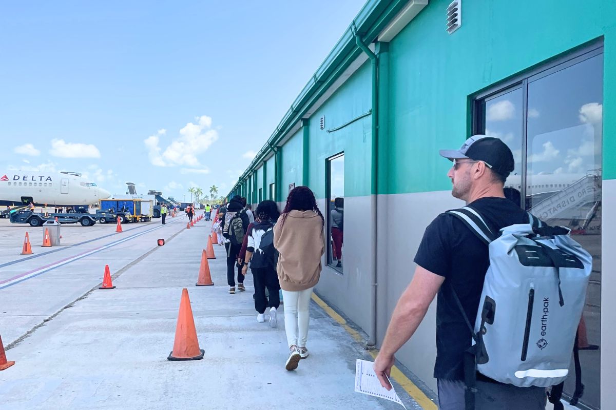 Passengers walking on the tarmac after landing at Belize City International Airport, with a Delta airplane in the background and a line of travelers heading towards the terminal building under a clear blue sky.