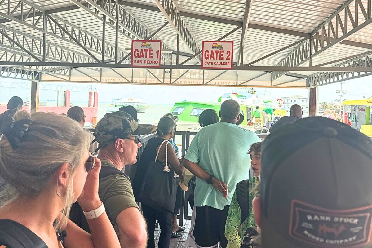 Passengers waiting to board the water taxi in Belize City, with signs indicating Gate 1 for San Pedro and Gate 2 for Caye Caulker. The dock and water taxis are visible in the background.