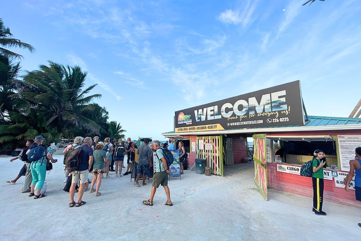 Travelers waiting for luggage at the water taxi terminal in Belize, with a "Welcome" sign and palm trees in the background.