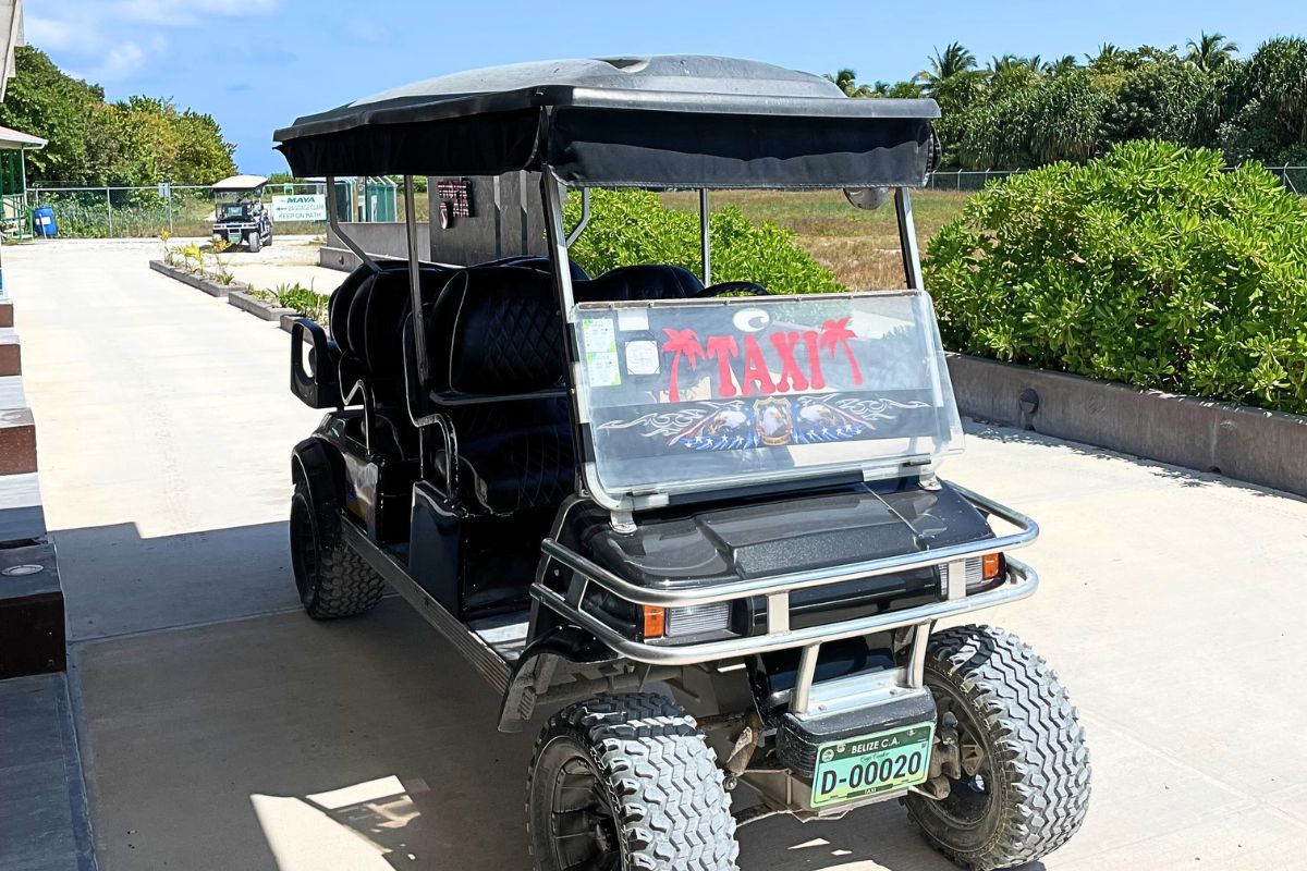 A golf cart taxi in Caye Caulker, Belize, with a "TAXI" sign on the windshield, parked on a road with greenery and a clear blue sky in the background.