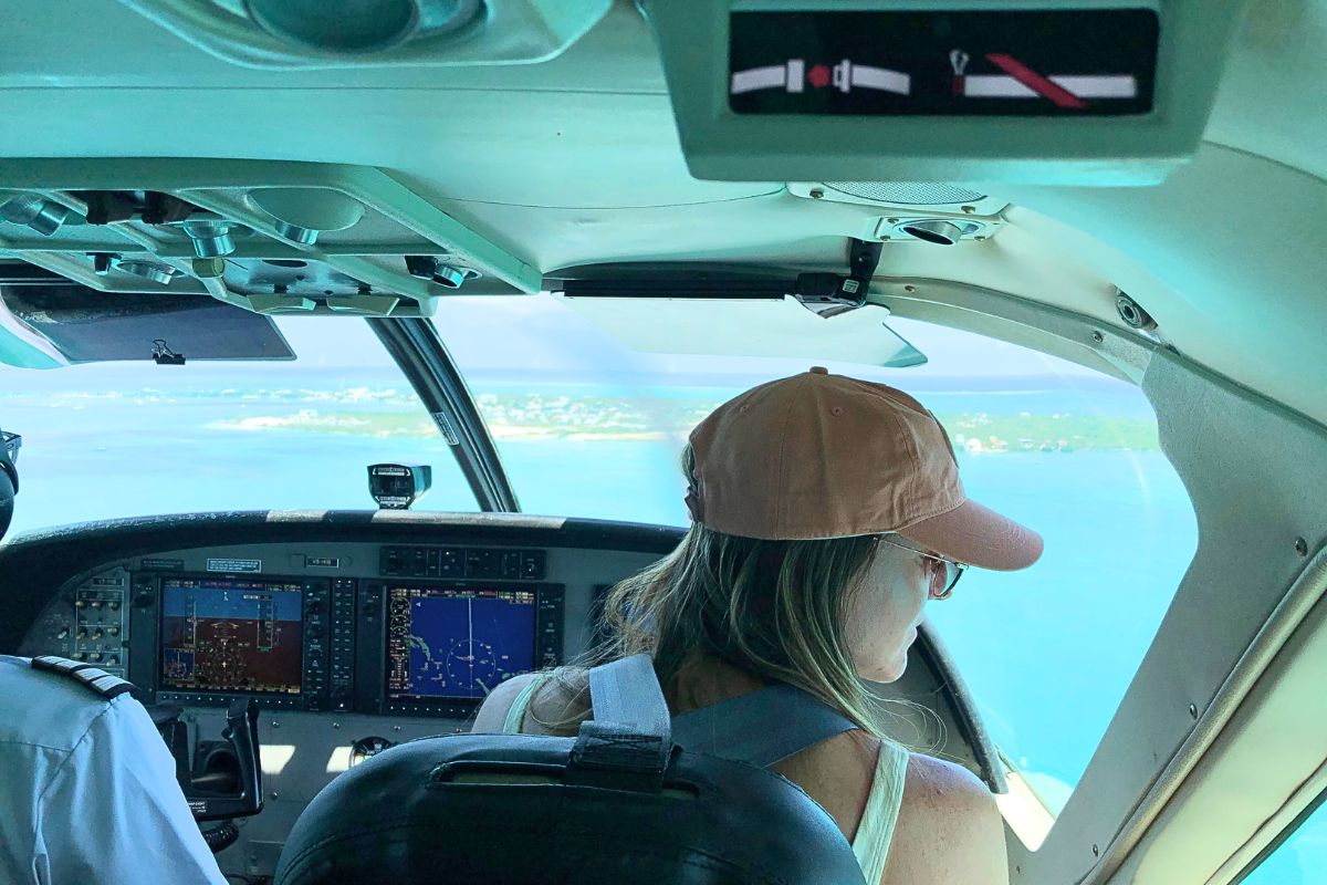  Kate sitting in the cockpit of a small plane, flying to the islands of Belize. The view through the windshield shows the turquoise waters and islands below. The pilot's controls and instruments are visible in the foreground.