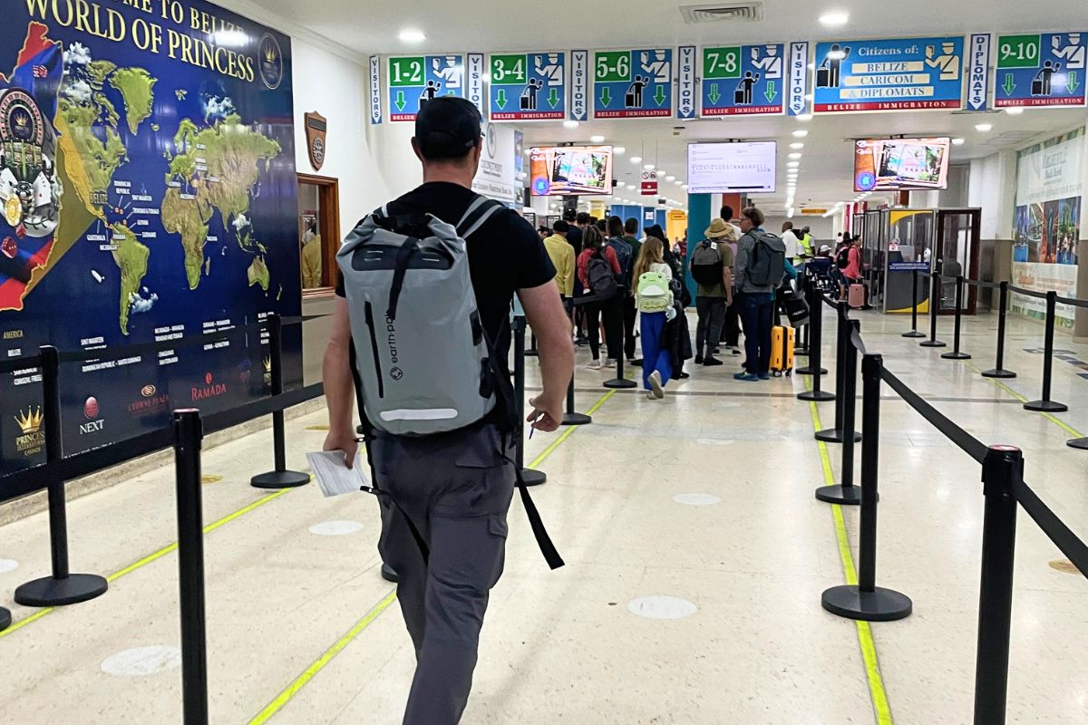 Travelers stand in line at the customs area of Belize City International Airport, where overhead signs indicate different lanes for visitors and citizens.