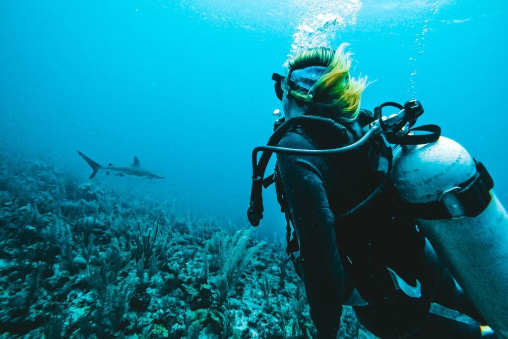 Kate scuba diving in Belize, with a shark in the distance, surrounded by underwater coral and marine life.