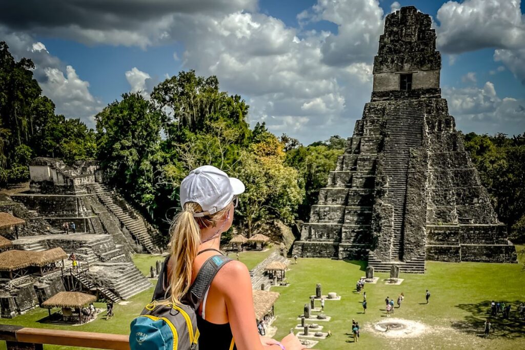 Kate exploring the ancient Mayan ruins at Tikal, Guatemala, wearing a white cap and backpack, with a view of the towering pyramid and surrounding structures.