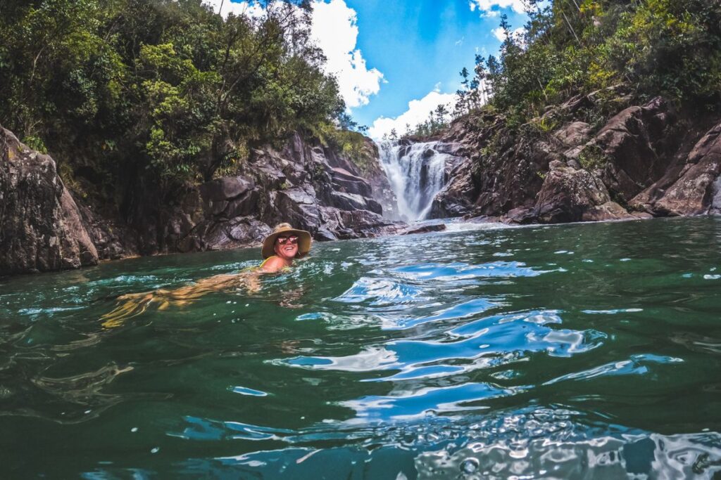 Kate swimming in the pool at the base of Big Rock Falls in Belize, wearing a sunhat and sunglasses, with the waterfall and lush greenery in the background.