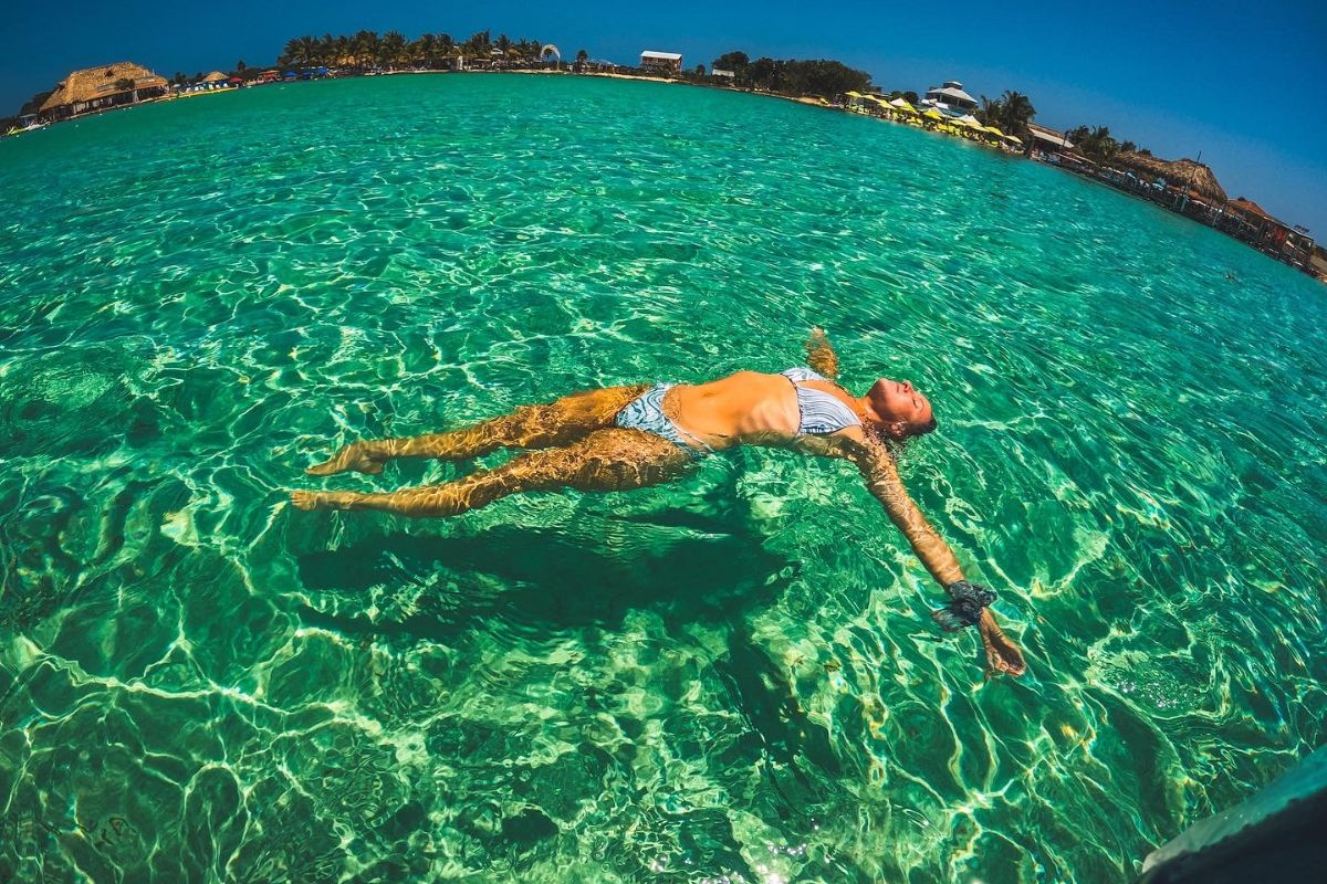 Kate floating on her back in the clear turquoise waters at Secret Beach on Ambergris Caye, Belize, with a view of the shoreline and palm trees in the distance, as featured in a Belize Travel Guide