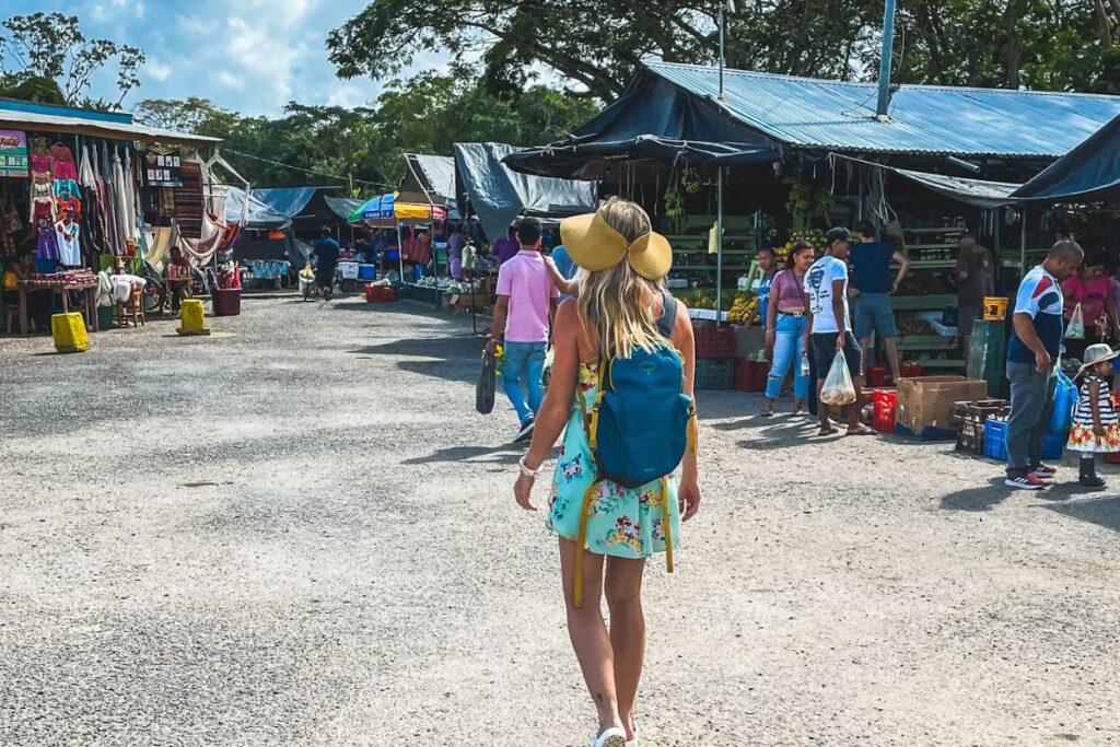 Kate walking through the farmers market in San Ignacio, Belize, wearing a floral dress and backpack, with market stalls and people in the background.