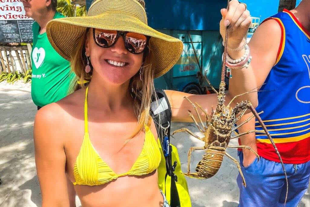 Kate enjoying lobster season in Belize, wearing a yellow bikini and sunhat, holding a large lobster with a smile on her face