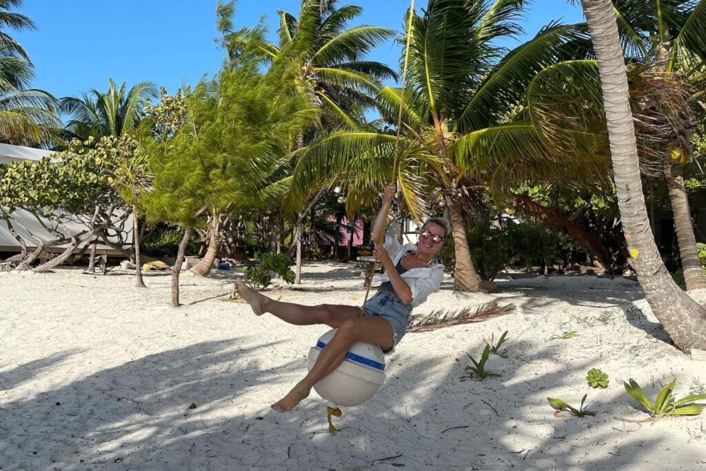 Kate having fun on a beach swing made from a buoy in Belize, surrounded by palm trees and sandy beach.