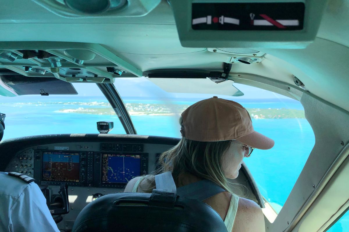 Kate is sitting in the cockpit of a small plane, flying over the turquoise waters of Belize, with islands visible through the plane windows. 