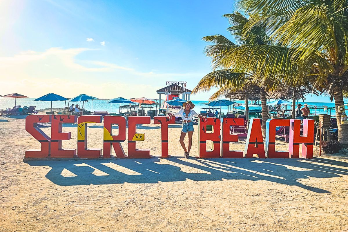 Kate posing by a large, colorful "Secret Beach" sign on a sandy beach in Belize. She is wearing a white top, denim shorts, and a wide-brimmed sun hat, embodying a relaxed, beachy vibe. The background features palm trees, beach umbrellas, and a clear blue sky.