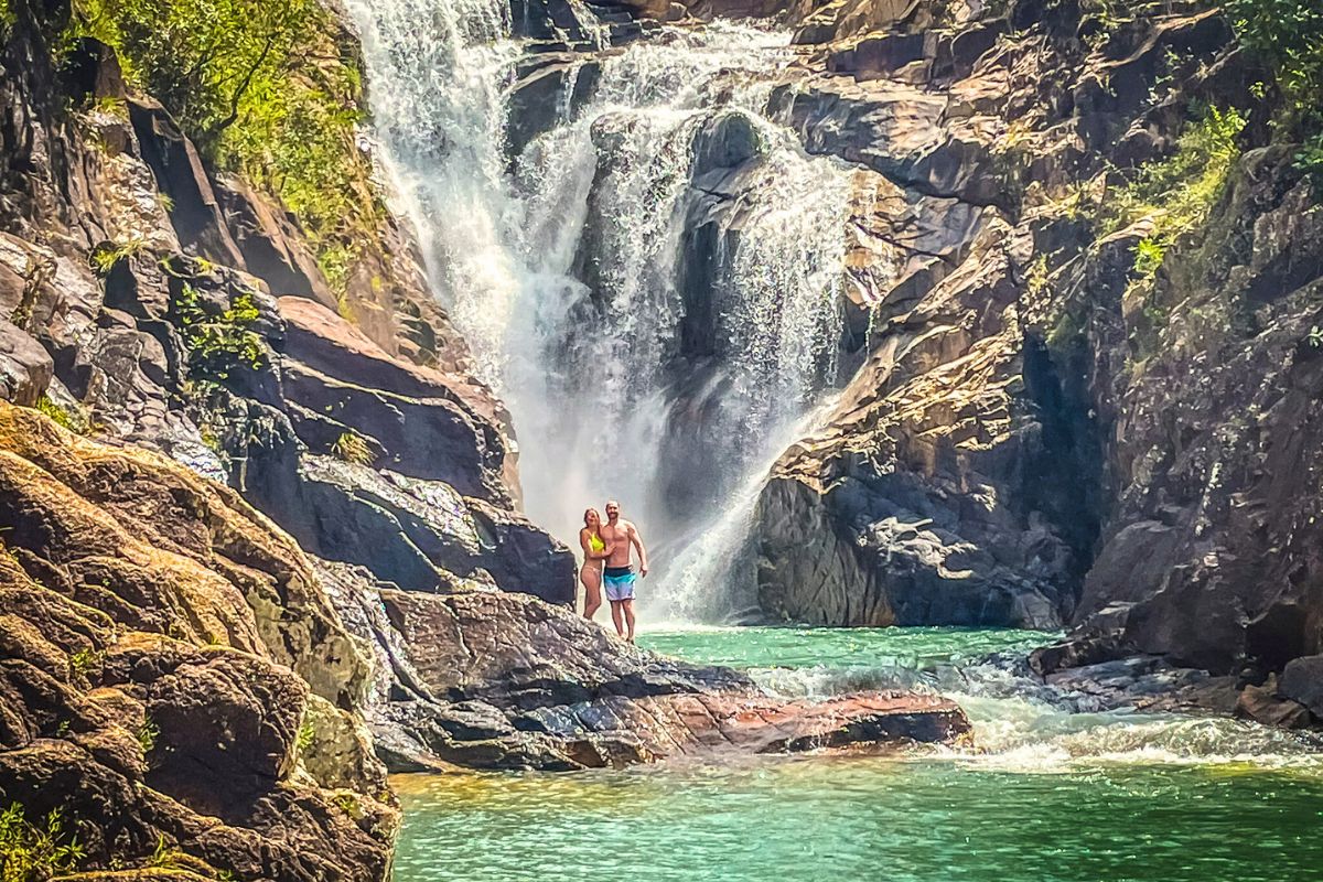 Kate and her husband standing together on a rocky outcrop at the base of a powerful waterfall named Big Rock Falls, in Belize. Kate is wearing a bright yellow bikini while her husband is in swim trunks. 