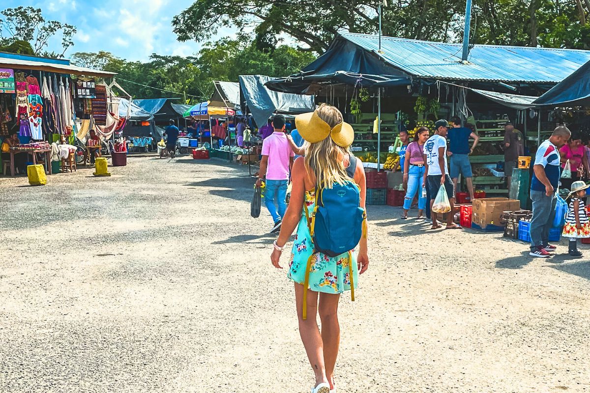 Kate explores a bustling local market in San Ignacio Belize, wearing a sunhat and a blue backpack. She walks past colorful stalls displaying handmade crafts, fresh produce, and vibrant clothing. The market is lively with people shopping and interacting, capturing the essence of local culture.