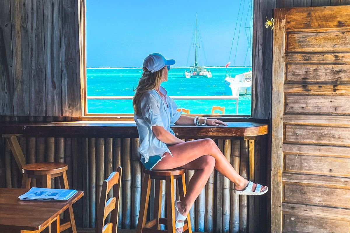 Kate sitting by a window in Belize, looking out at the turquoise waters and sailboats. She is wearing a white shirt, denim shorts, and a white cap, enjoying the beautiful view and relaxed atmosphere.