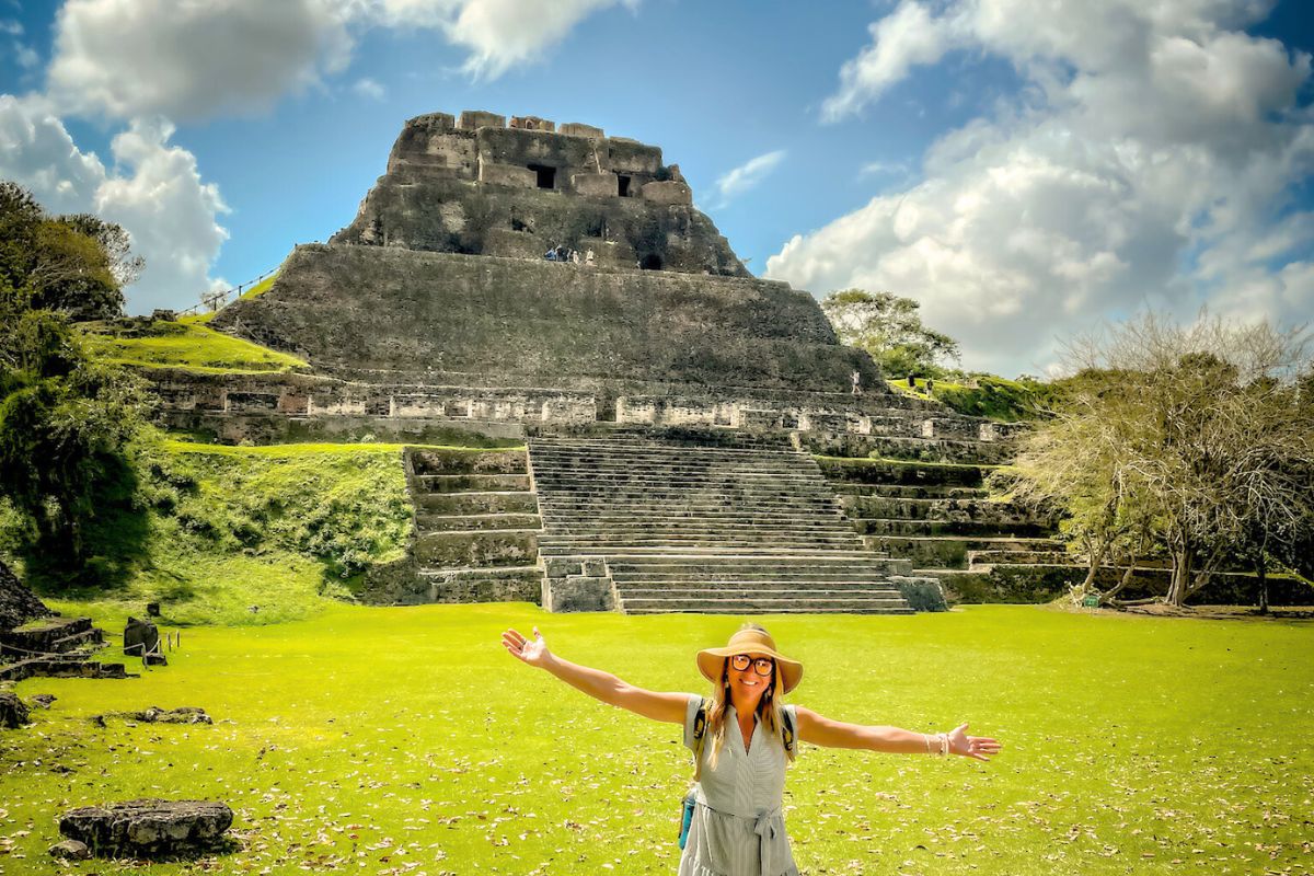 Kate standing in front of an ancient Mayan pyramid in Belize, arms outstretched and smiling. She is wearing a sun hat and a light dress, enjoying the historical site and lush green surroundings on a bright day.