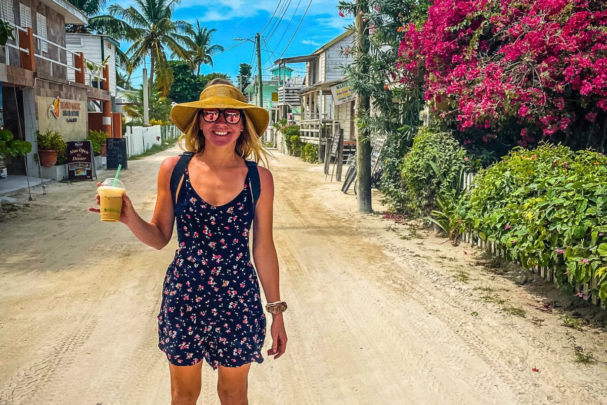 Kate walking down a sandy street on Caye Caulker, Belize, holding a cold drink. She is wearing a floral dress, sun hat, and sunglasses, with tropical plants and colorful buildings lining the street.