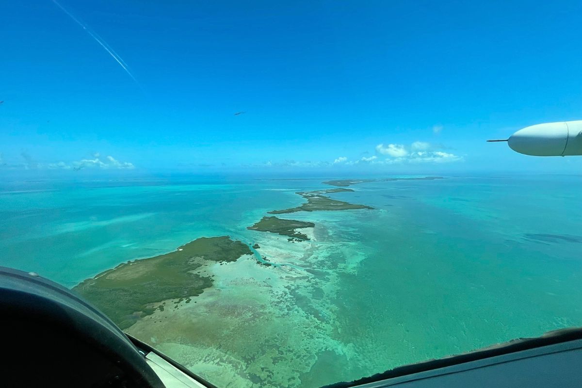 Aerial view of the turquoise waters and small islands of Belize, taken from a plane flying over the Caribbean Sea. The clear blue sky stretches out above the vibrant, shallow waters.