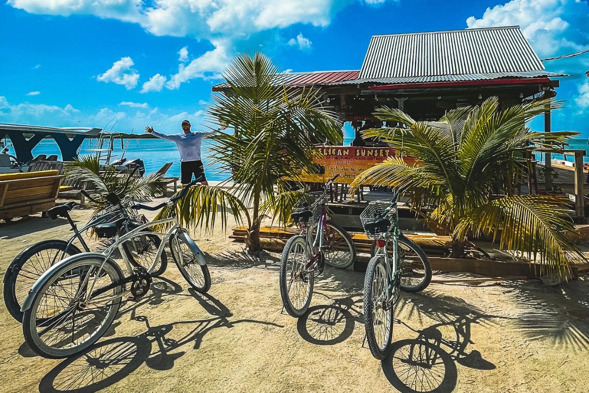 Bicycles parked outside a small beachside bar called Pelican Sunset Bar in Caye Caulker, Belize. A man stands with arms outstretched near the water, surrounded by palm trees and outdoor seating.