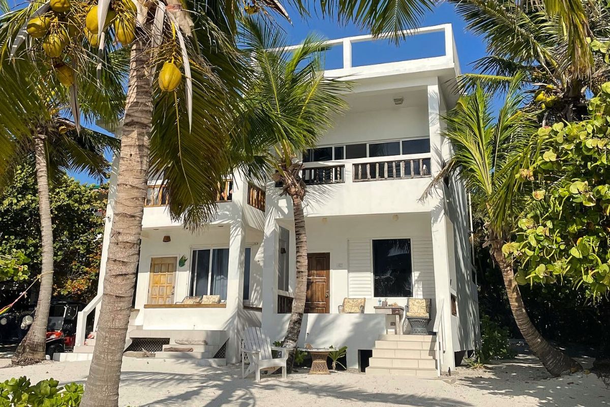 A two-story white beachfront house in Ambergris Caye, Belize, surrounded by palm trees and tropical vegetation. The house features balconies and a small patio area with outdoor seating.