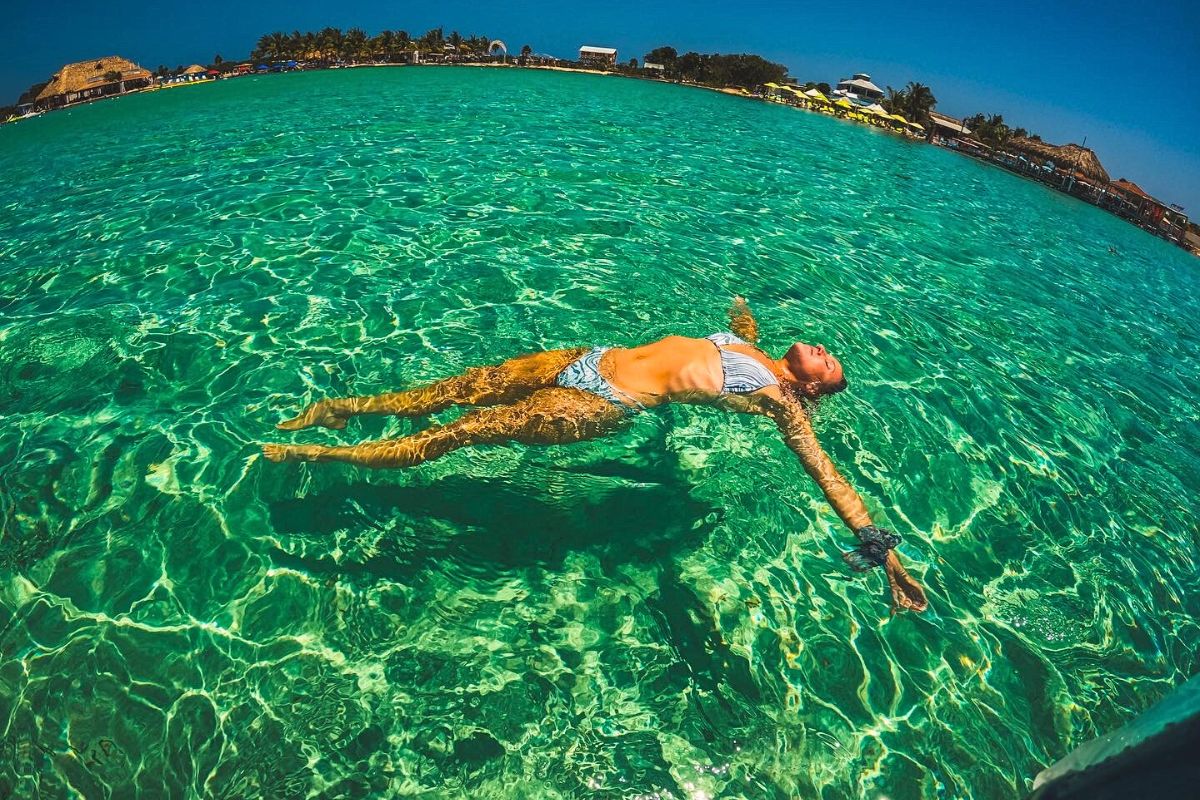 Kate from Kate's Crossing Blog floating peacefully in the clear turquoise water off Ambergris Caye, Belize. She is wearing a bikini and enjoying the sunny day, with the shoreline and palm trees visible in the distance.