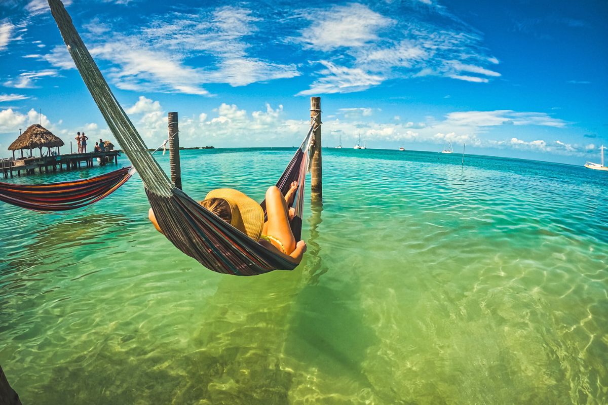 Kate from Kate's Crossing Blog relaxing in a hammock suspended over the clear turquoise water in Caye Caulker, Belize. She is wearing a sun hat, with sailboats and a thatched-roof pier visible in the background.