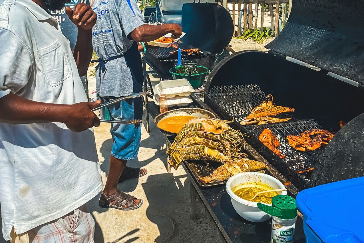 Street food vendors grilling fresh seafood on Caye Caulker, Belize. Lobsters and fish are being cooked on large grills, with a variety of sauces and seasonings in bowls nearby.