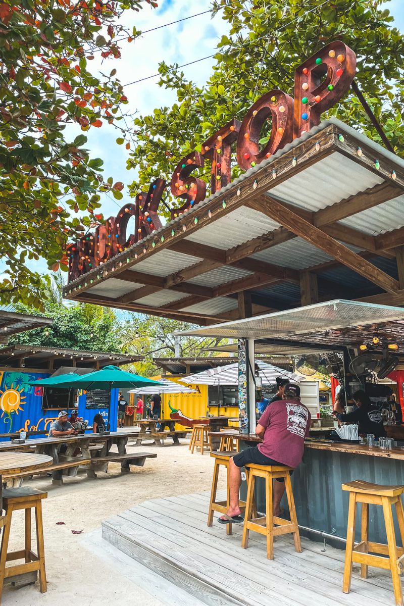 Food trucks at The Truck Stop on Ambergris Caye, Belize. The area features colorful containers converted into food stalls, picnic tables, and a bar with stools, all under the shade of trees decorated with lights.