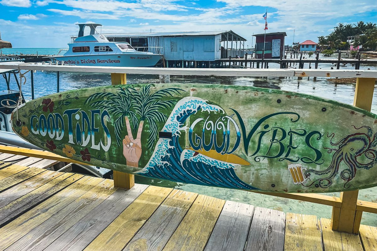 A colorful surfboard sign with the text 'Good Tides, Good Vibes' displayed on a dock in Caye Caulker, Belize. In the background, boats and waterfront buildings are visible against the blue sky and ocean.