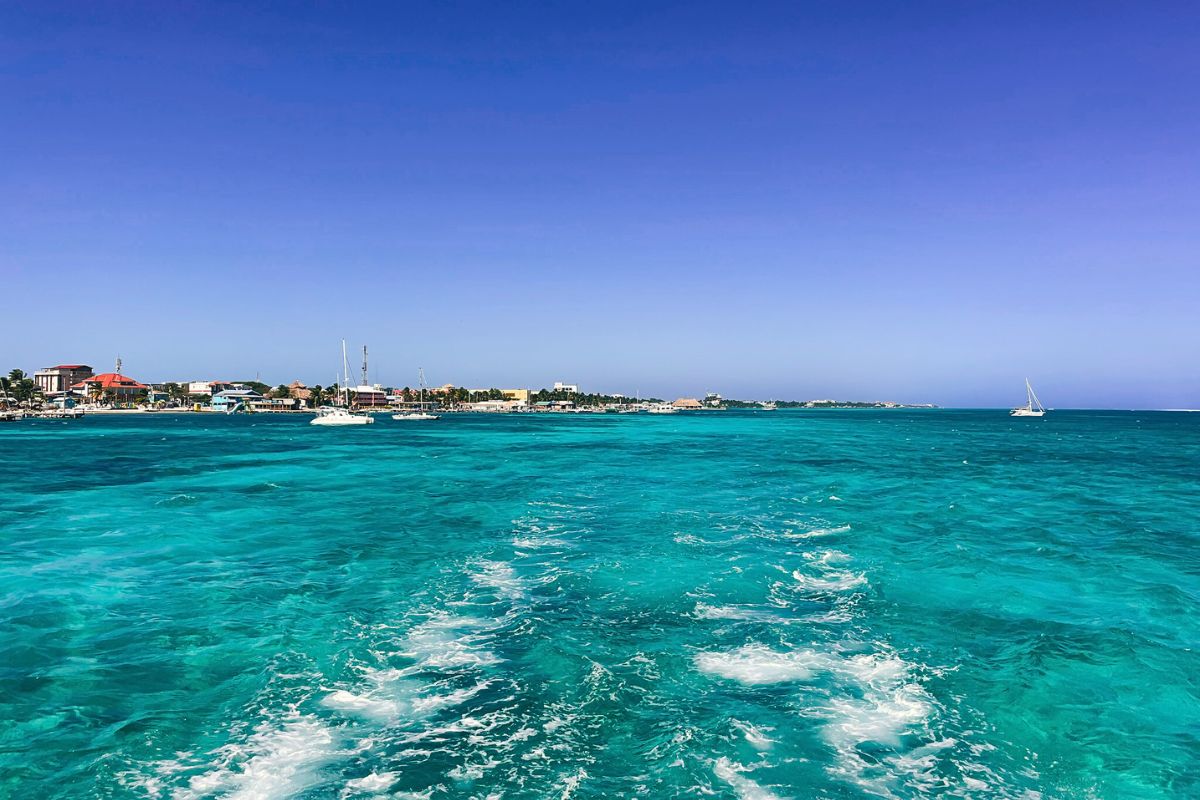 View from a water taxi between Caye Caulker and Ambergris Caye, Belize. The image shows the vibrant turquoise ocean with small boats and the shoreline of a coastal town in the distance under a clear blue sky.