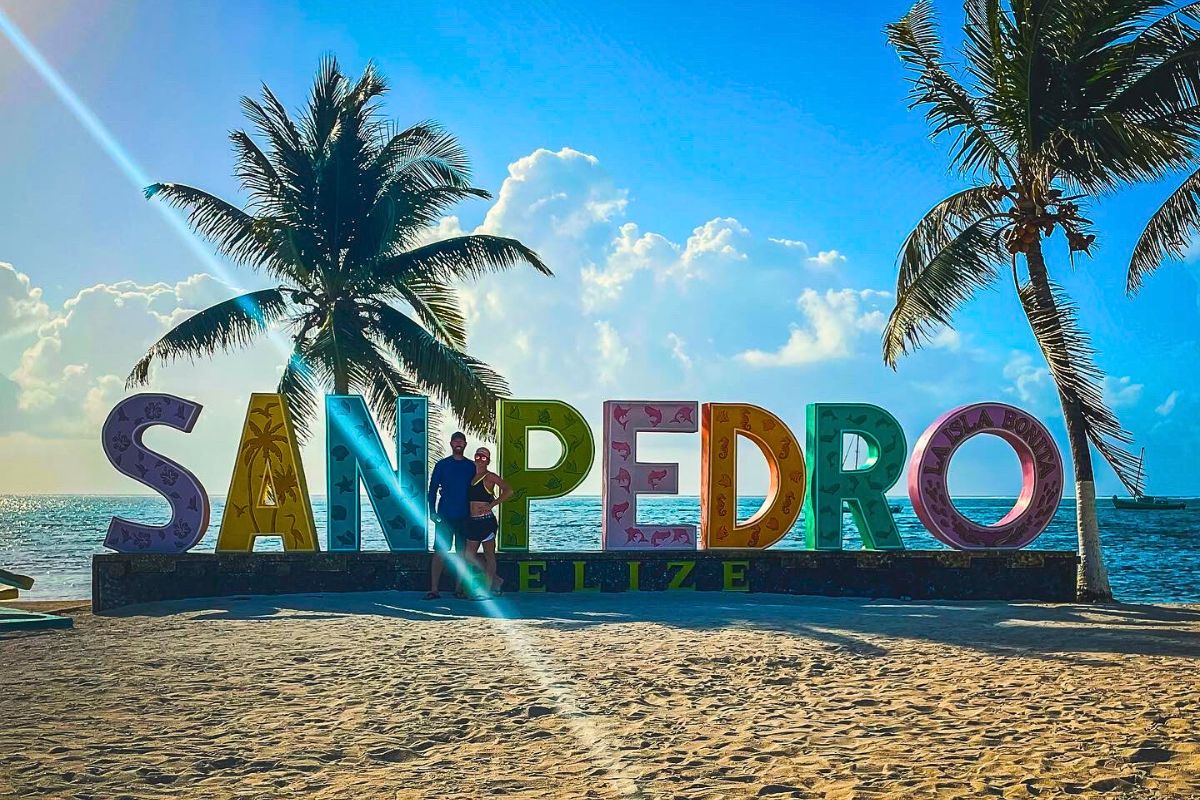 A large 'San Pedro' sign with colorful letters stands on a sandy beach, flanked by palm trees. Kate from Kate's Crossing Blog and her husband pose in front of the sign with the ocean and a clear blue sky in the background.