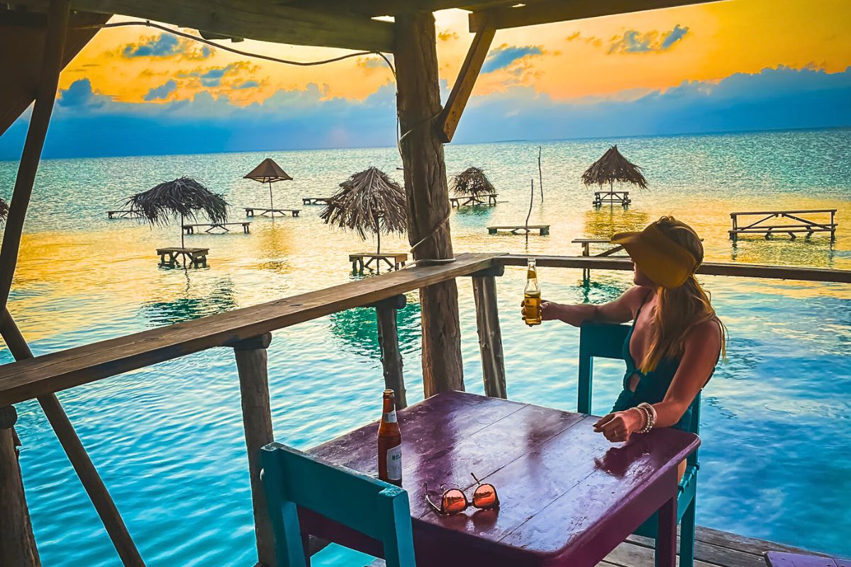 Kate enjoying a drink at a waterfront bar on Ambergris Caye, Belize, during sunset. She is seated at a table overlooking the calm sea with thatched-roof picnic tables in the water, reflecting the vibrant colors of the sunset.