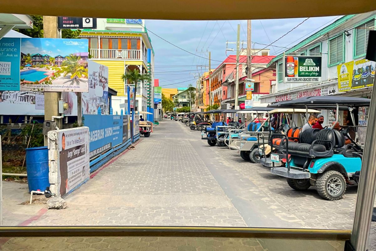 A street in Ambergris Caye, Belize, lined with colorful buildings and parked golf carts. Signs advertise local businesses, including a grocery store. People walk and drive golf carts along the road.