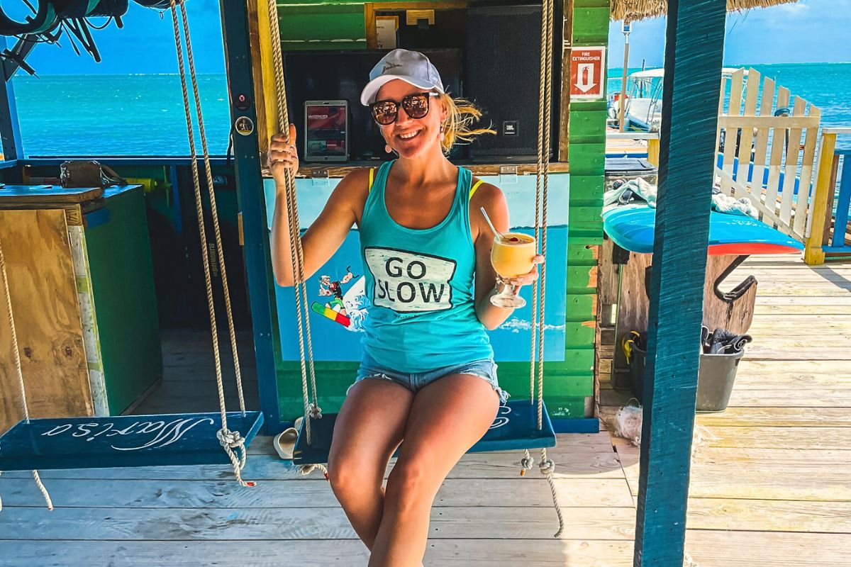 Kate sitting on a swing at a beachside bar in Caye Caulker, Belize, wearing a 'Go Slow' tank top and holding a tropical drink. The turquoise ocean and colorful bar decor create a relaxing atmosphere.