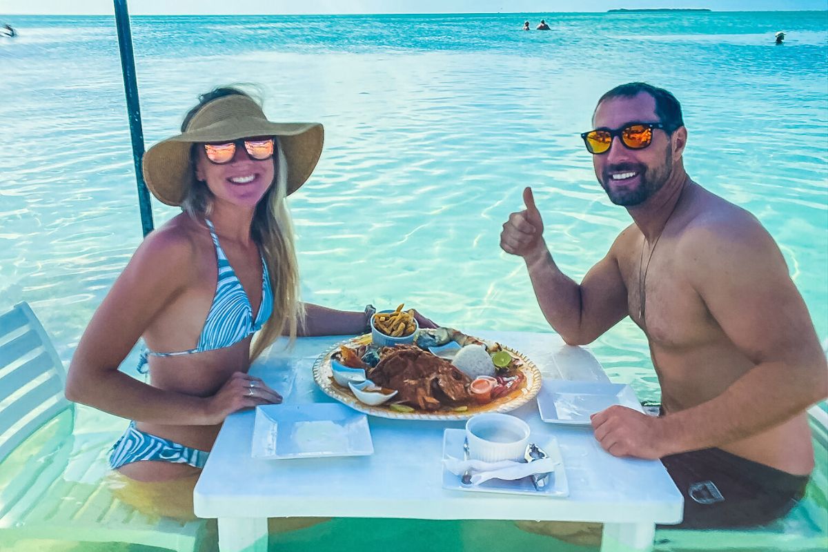 Kate and her husband dining at a table set in shallow ocean water in Ambergris Caye, Belize. Both are in swimwear and sunglasses, enjoying a seafood meal with the turquoise sea stretching out behind them.