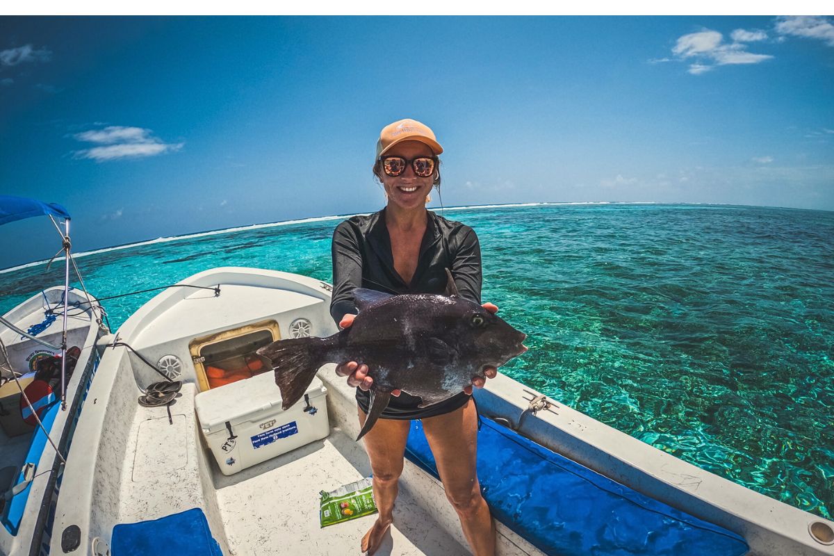 Kate on a boat in Caye Caulker, Belize, holding a large fish she caught. She is smiling, wearing sunglasses and a cap, with the clear blue ocean in the background.