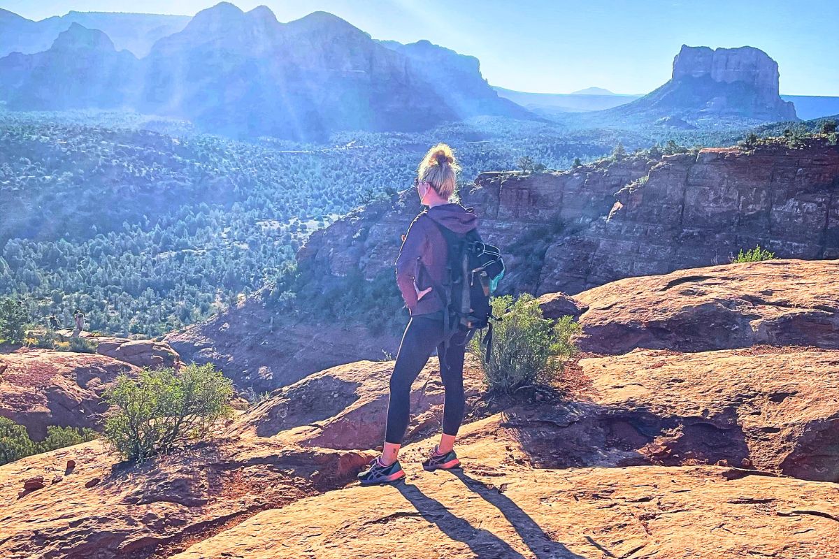 Kate hiking the Cathedral Rock Trail in Sedona, standing on a red rock ledge, looking out over a stunning landscape of green valleys and rugged cliffs under a clear blue sky.