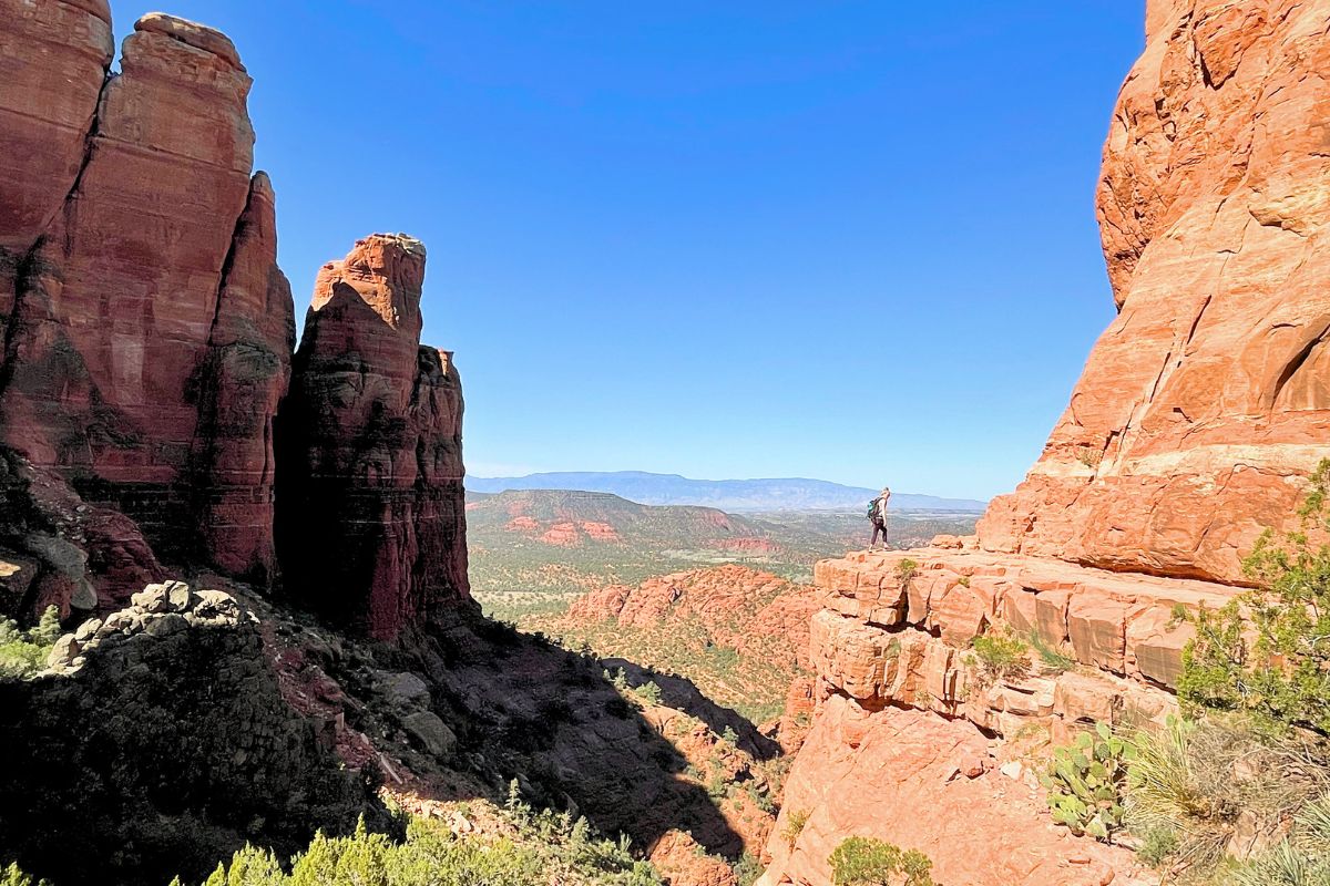 Kate standing on a rocky ledge at the top of Cathedral Rock in Sedona, surrounded by panoramic views of red rock formations and valleys under a clear blue sky.