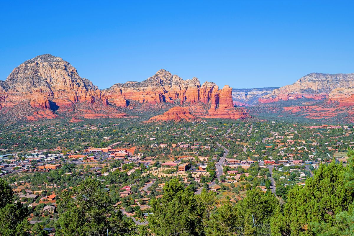 Panoramic view of Sedona, showcasing the town's layout with its iconic red rock formations and mountains in the background under a clear blue sky.