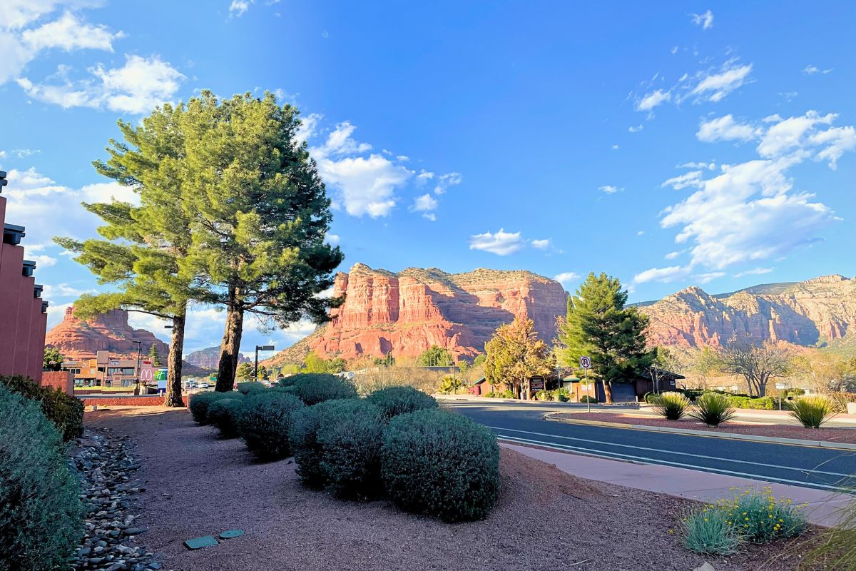 Street view in Sedona, featuring the red rock formations in the background, with a clear blue sky and scattered clouds above.