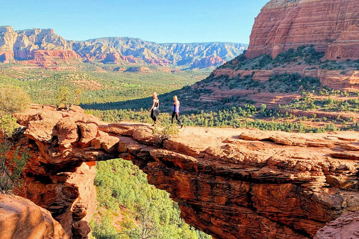 Two hikers walking across Devil's Bridge in Sedona, with expansive views of the red rock formations and valleys in the background under a clear blue sky.