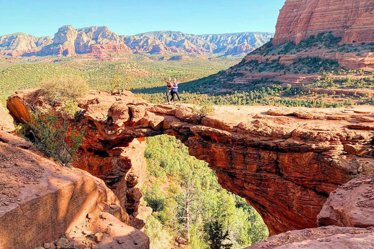 Kate from Kate's Crossing Blog and a friend standing on Devil's Bridge in Sedona, with stunning red rock formations and lush green valleys stretching out behind them.